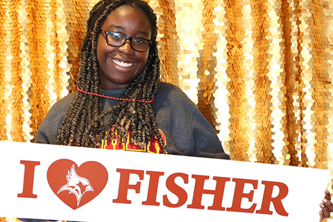 A smiling person holds a sign reading I HEART FISHER against a gold sequin backdrop, conveying a proud, cheerful emotion.